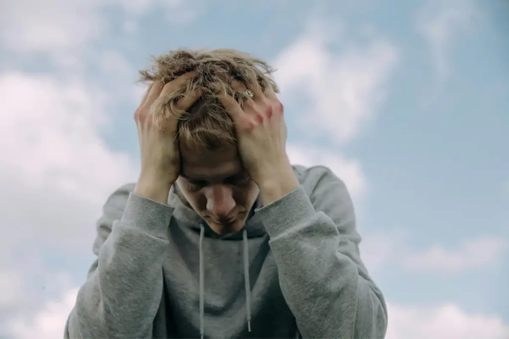 Young man in gray hoodie holds head in frustration, set against cloudy sky