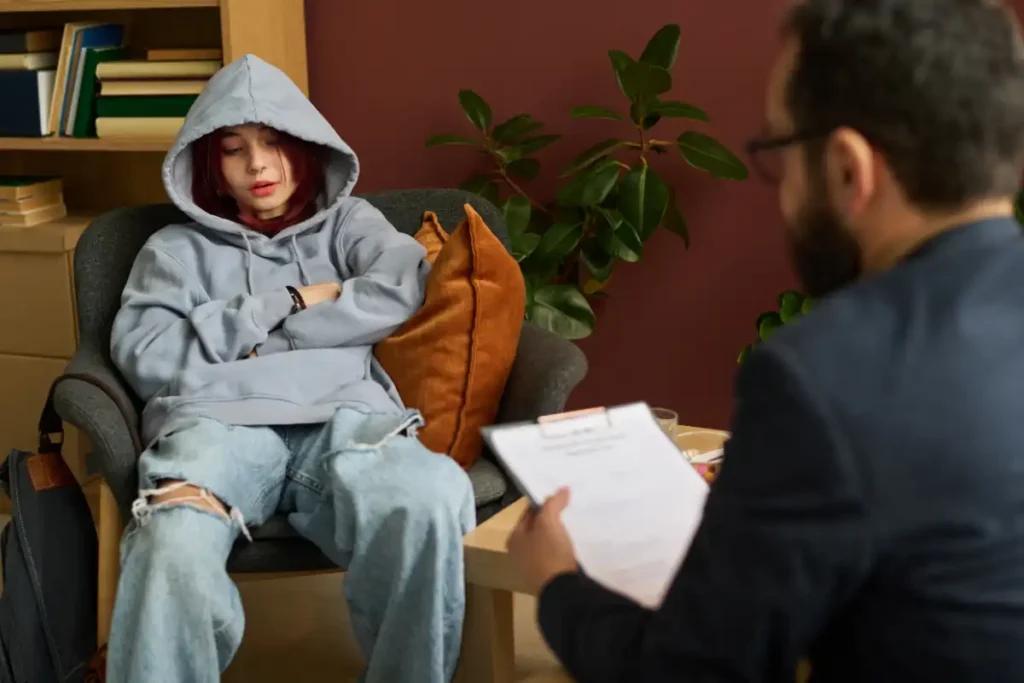 Youthful girl in casual attire describing her problem to school psychologist with document sitting in front of patient